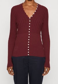 Viininpunainen resorineuloksinen neuletakki V-kaula-aukolla, jossa valkoiset napit edessä ja aaltoilevat reunat, pehmeästä neulosmateriaalista valmistettu.