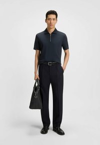 Marinblå kortärmad polo med dragkedjekrage, parat med skräddarsydda mörka byxor. Modellen håller en svart texturerad handväska och bär svarta slip-on skor.