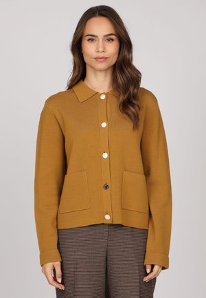 Femme portant un cardigan à boutons jaune moutarde avec deux poches avant et un pantalon à carreaux marron, se tenant devant un fond uni.