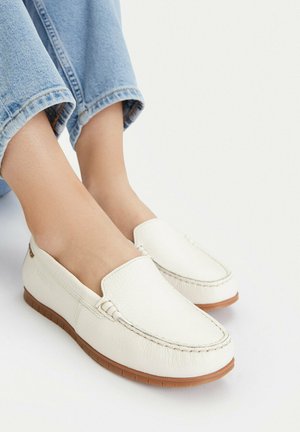 Mocassini bianchi in pelle senza lacci con suole di gomma marroni indossati con jeans azzurri su uno sfondo bianco.