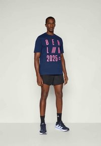 Navy blue cotton t-shirt featuring pink graphic text "BERLIN 2025" and stripes. Paired with black shorts and navy athletic shoes.