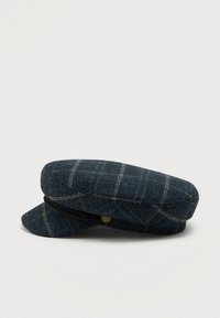 Sininen tweed-tyylinen lippalakki, jossa on vaaleanruskea ikkunaruudullinen kuvio, musta punottu nauha ja pieni kultainen embleemi sivulla, asetettuna tasaiselle vaalealle pinnalle.