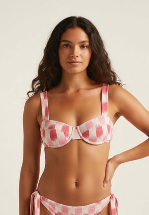 Mujer con cabello largo y rizado vistiendo un top de bikini a cuadros rosa y blanco y braguitas a juego sobre un fondo liso.