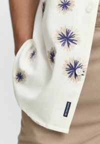 Weißes Stoffhemd mit gestickten blauen und beigen Blumenmustern, mit Knopfleiste und einem Etikett. Weicher Textil.