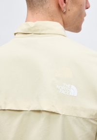 Άτομο που φοράει ένα ανοιχτό μπεζ μπουφάν North Face, ορατό από πίσω και πλευρά, με ένα μικρό σκουλαρίκι κρίκο στο αριστερό αυτί.