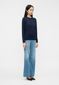 Μπλε πουλόβερ με μακριά μανίκια και λεπτομέρειες με κουμπιά στα μανίκια, συνδυασμένο με ανοιχτό μπλε τζιν παντελόνι φαρδύ στο κάτω μέρος. Μαύρα μυτερά παπούτσια ολοκληρώνουν την εμφάνιση.