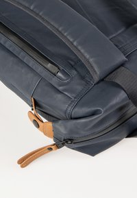 Zaino in pelle blu navy con cerniere nere, dettagli in pelle beige e una superficie liscia; presenta una tasca laterale con chiusura a fibbia.