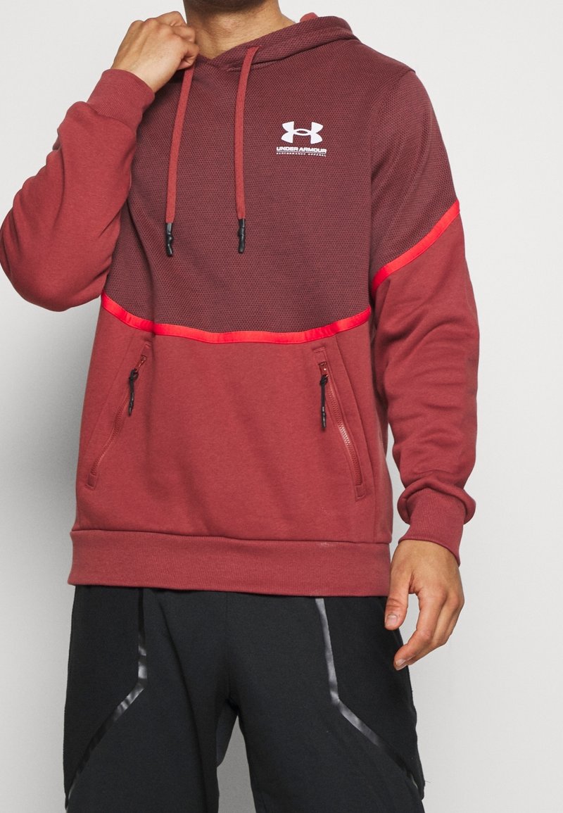 Rote Kapuzenjacke mit strukturiertem oberen Abschnitt, schwarzen Reißverschlusstaschen und Kordeln. Verfügt über reflektierende Akzente und Logo auf der Brust.