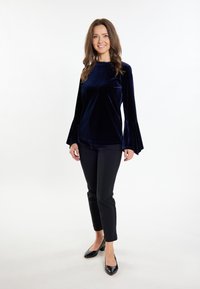Top in velluto blu navy con maniche a campana, abbinato a pantaloni neri slim e décolleté nere a punta, su uno sfondo bianco.
