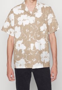 Camicia a maniche corte beige con motivo floreale bianco, realizzata in tessuto leggero. Presenta una chiusura con bottoni sul davanti e una tasca singola sul petto.