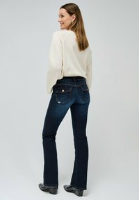 Jeans de campana azul oscuro con cintura de altura media, combinados con un suéter color crema. Presentan bolsillos traseros y una sutil textura de mezclilla.