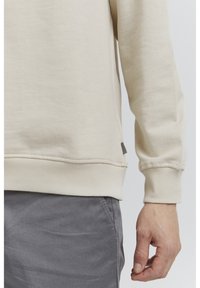 Sudadera beige con dobladillo y puños acanalados. Presenta una etiqueta lateral y una textura suave. Combinada con pantalones grises.