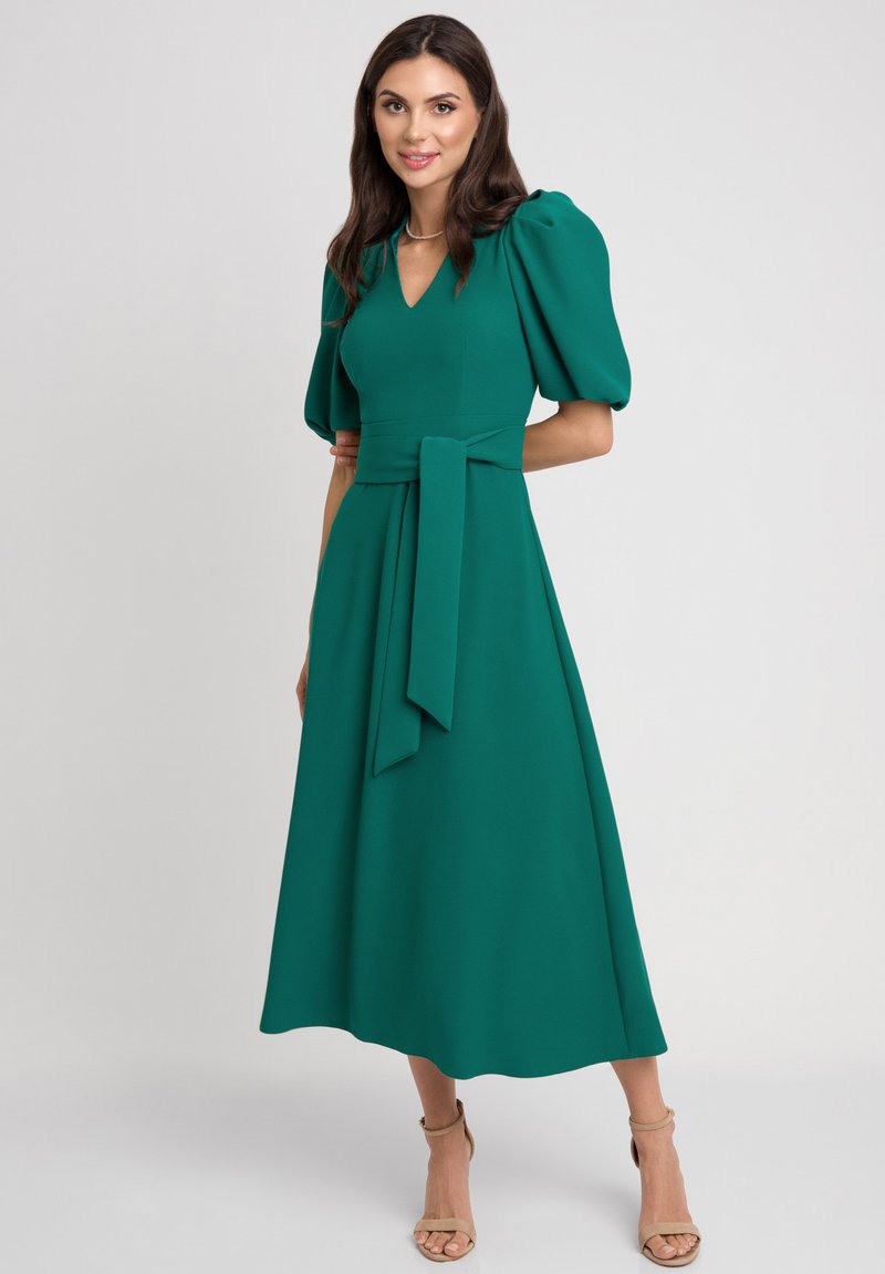 Zielona midi sukienka z bufiastymi rękawami, dekoltem w kształcie litery V i związywanym paskiem w talii. Wykonana z gładkiego materiału, posiada zwiewną spódnicę i neutralne obcasy na kostkę.