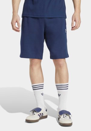 Marineblauwe sportshorts met een elastische tailleband, voorzien van zijzakken en een klein wit logo. Gepaarde met gestreepte witte sokken en sneakers.