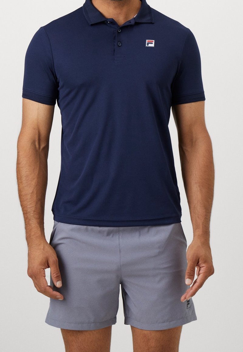 Homme portant un polo bleu marine à manches courtes et un short gris clair, debout devant un fond uni.