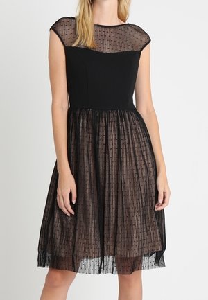 Kobieta w czarnej sukience do kolan z przezroczystym materiałem w groszki na ramionach i spódnicy, dopasowana w talii.