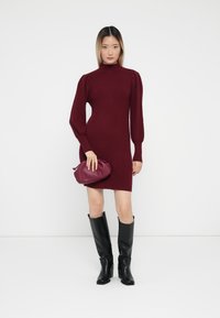 Burgundy ribbed knit dress with a high neck and puff sleeves, paired with knee-high black boots and a small burgundy handbag.