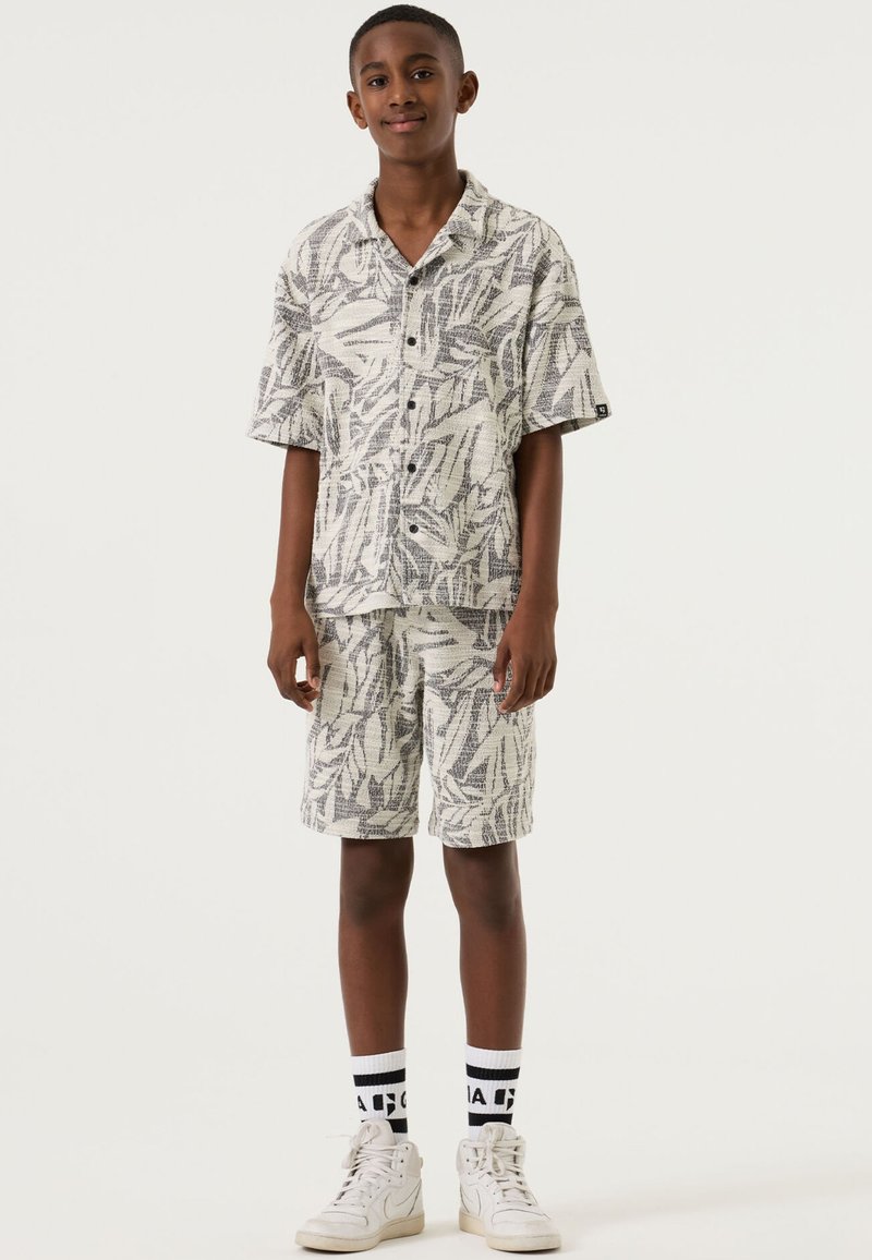 Camicia a maniche corte con bottoni e pantaloncini abbinati con motivo a foglie in bianco e nero, realizzati in tessuto leggero. Scarpe da ginnastica bianche e calzini con fantasia.