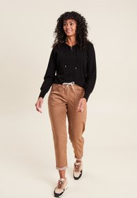 Zwart hoodie met trekkoorden, tan broek met opgerolde manchetten en beige sneakers met donkere accenten. Model loopt tegen een neutrale achtergrond.