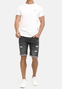 Camiseta blanca de manga corta con un pequeño logo, combinada con pantalones cortos de mezclilla oscuros con detalles rasgados y puños doblados, complementada con zapatillas blancas.