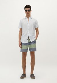Chemise à manches courtes bleu clair avec des rayures subtiles, associée à un short de bain à motifs vert et gris, et des espadrilles grises.