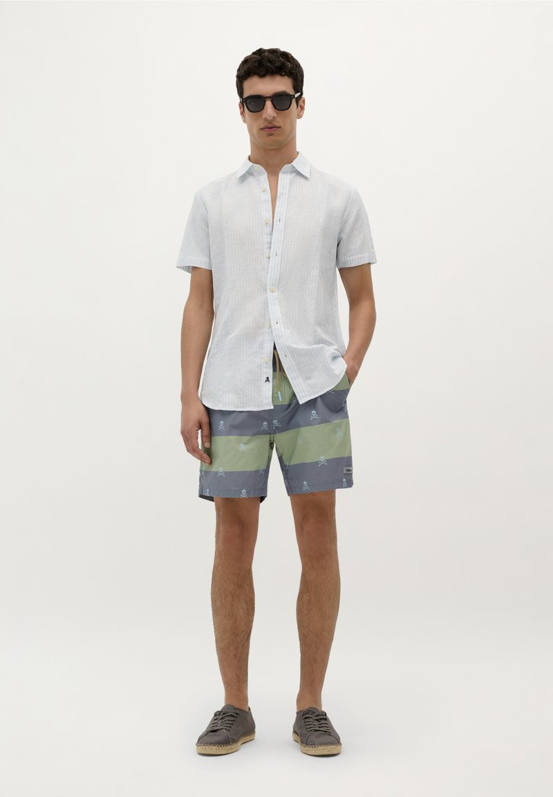 Chemise à manches courtes bleu clair avec des rayures subtiles, associée à un short de bain à motifs vert et gris, et des espadrilles grises.