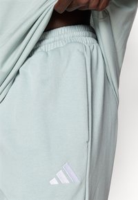 Gaismi zaļas sporta bikses ar elastīgu vidukļa joslu, uz kurām pie kabatas ir balts izšūts adidas logo, un mīkstu, gludu tekstūru.