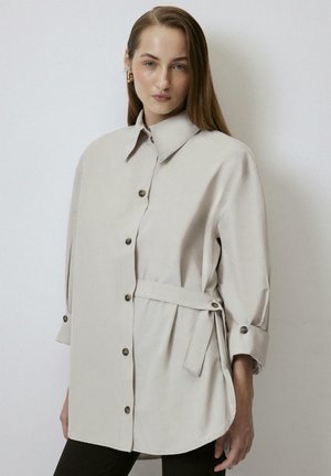 Camisa de botón de gran tamaño color beige claro con cuello alto, mangas largas, cinturón de lazo lateral y detalle de dobladillo redondeado. Textura de tela suave.