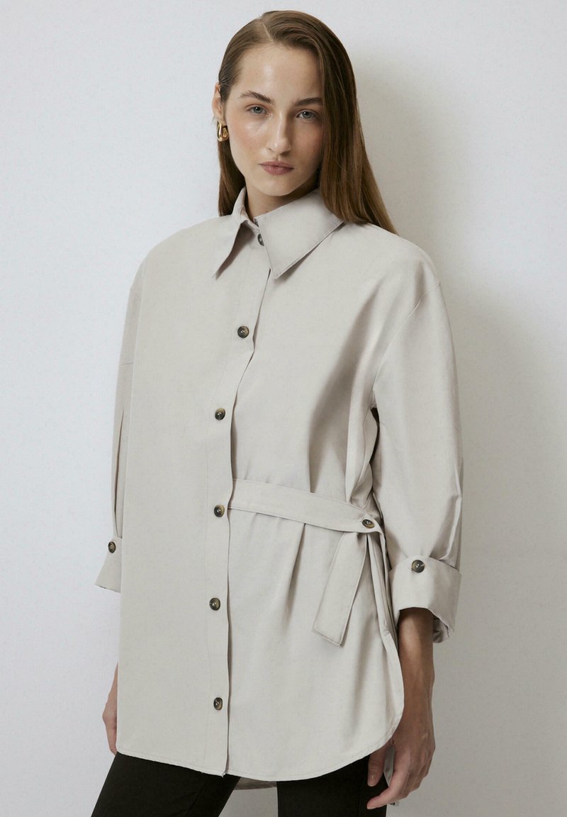 Camicia button-up oversize di un beige chiaro con colletto alto, maniche lunghe, cintura laterale e dettaglio dell'orlo arrotondato. Tessuto morbido.