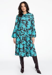 Ein türkises Blumenkleid mit schwarzem Hintergrund, Knopfverschluss, Kragen und Glockenärmeln, kombiniert mit knielangen, glänzenden burgunderroten Stiefeln.