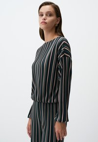 Maglietta a maniche lunghe a righe in verde scuro, caratterizzata da strisce verticali in diverse tonalità di beige, marrone e teal. Tessuto morbido, vestibilità comoda, scollatura a barca.