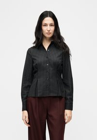 Γυναίκα με μακριά σκούρα μαλλιά, που φοράει μαύρο πουκάμισο με λεπτές ρίγες και σκούρο βιολετί παντελόνι, στέκεται μπροστά σε λευκό φόντο.