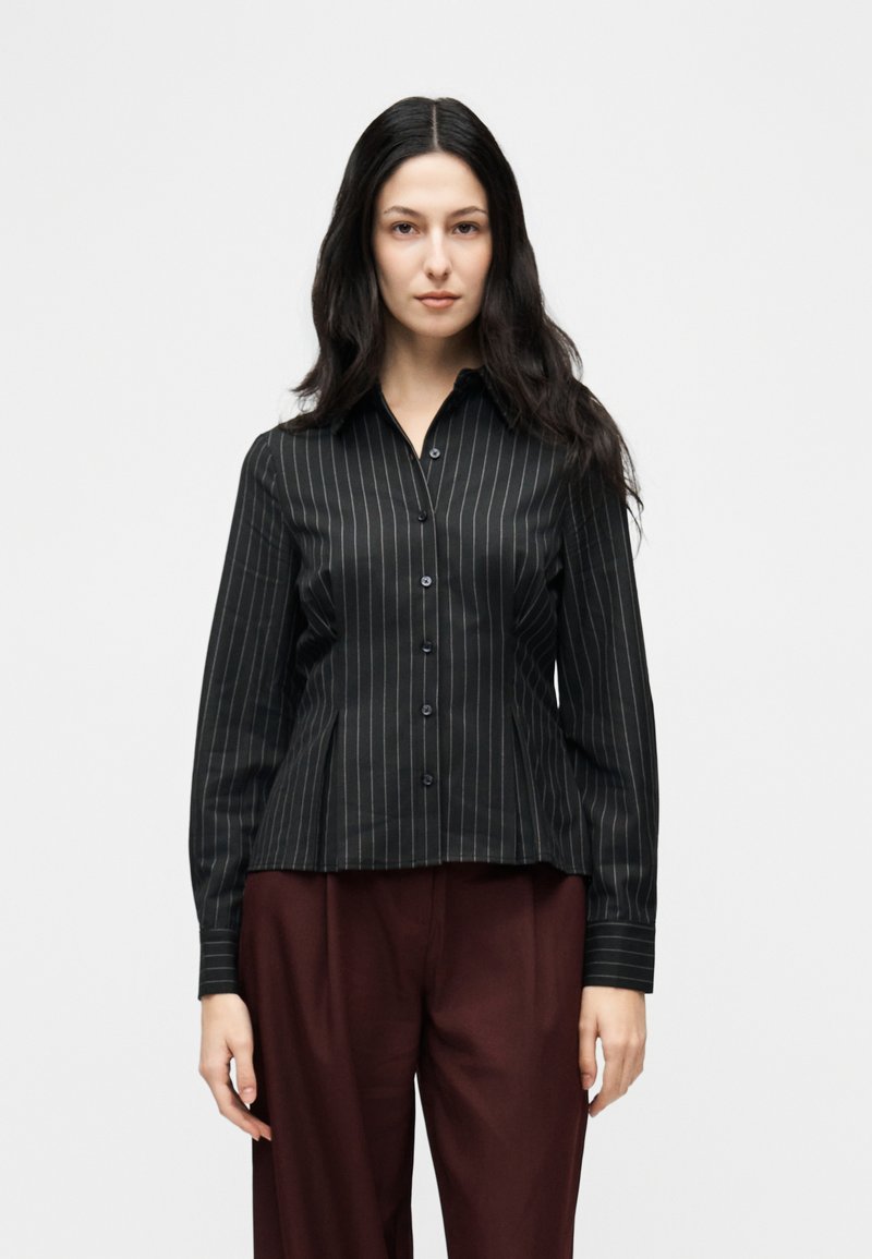 Γυναίκα με μακριά σκούρα μαλλιά, που φοράει μαύρο πουκάμισο με λεπτές ρίγες και σκούρο βιολετί παντελόνι, στέκεται μπροστά σε λευκό φόντο.