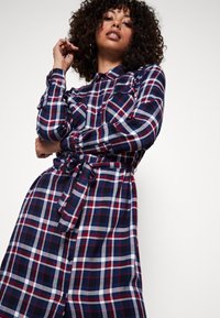 Abito camicia a quadri blu navy e rosso con colletto, maniche lunghe, chiusura frontale a bottoni e vita con cintura. Presenta due tasche sul petto.