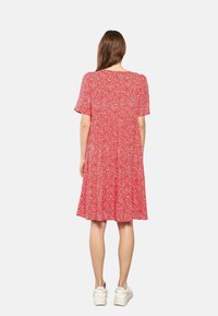 Femme aux longs cheveux bruns portant une robe rouge à fleurs arrivant au genou et des baskets blanches, se tenant de dos sur un fond uni.