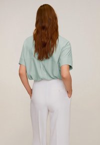 Personne avec de longs cheveux roux portant une chemise à manches courtes vert clair à l'intérieur d'un pantalon blanc taille haute, les mains dans les poches, de dos.