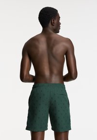 Žalios maudymosi šortai su tamsiai mėlynu kaktuso raštu, elastinga liemens juosta, šoniniais kišenėmis ir trumpa ilgio. Modelis parodytas iš nugaros.