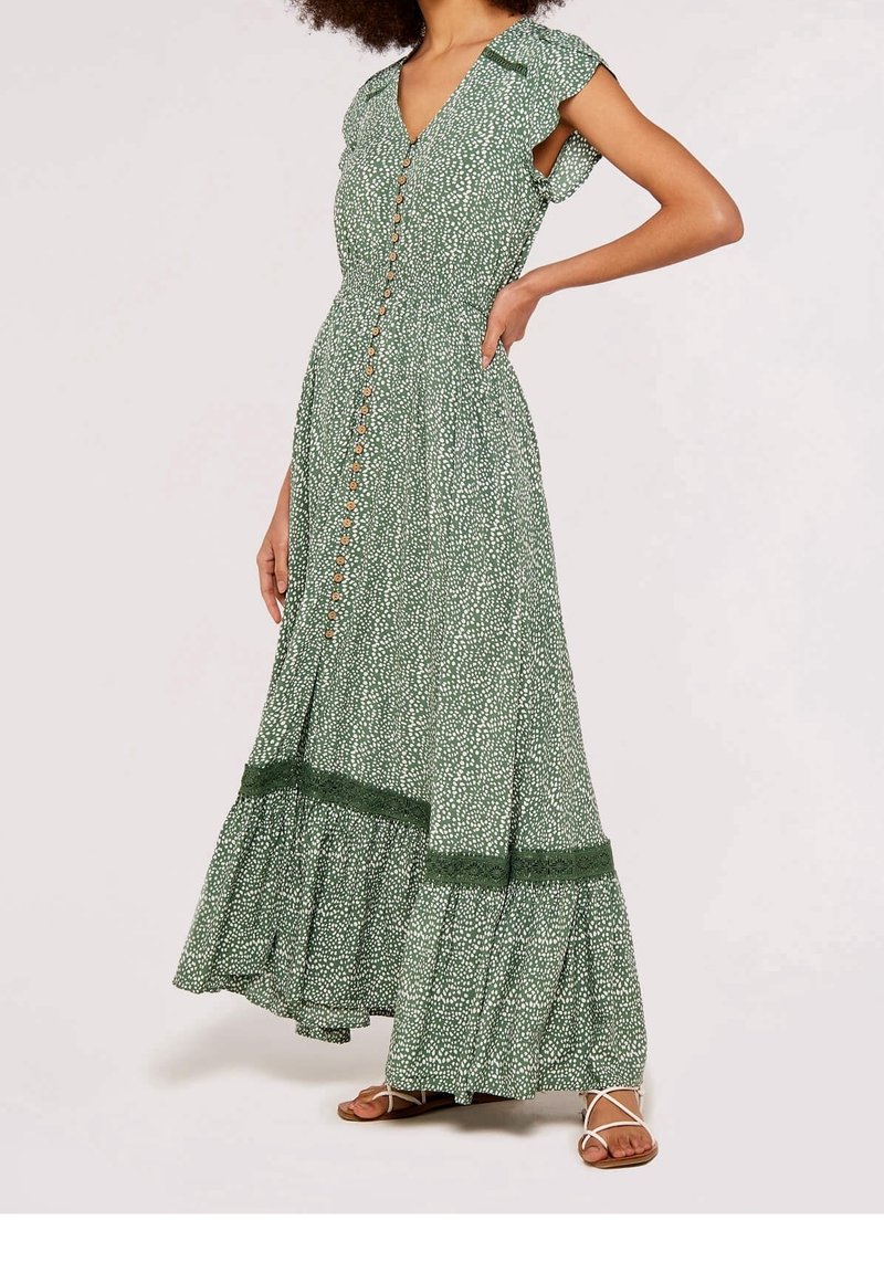 Femme portant une robe maxi sans manches à motif vert avec des boutons sur le devant et une bordure en dentelle près de l’ourlet, associée à des sandales blanches.
