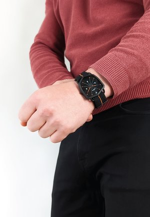 Sort rektangulær ur med orange accenter på et stofarmbånd, båret på et håndled. Sweater er bordeaux, og bukserne er sorte.