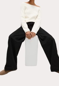 Personne assise sur un tabouret rectangulaire blanc, portant un pull blanc à épaules dénudées, un pantalon large noir et des chaussures marron, les mains reposant sur les genoux.