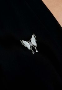 Silver butterfly brooch with pearlescent wings and a central stone, featuring two dangling crystal drops; set against a black background.