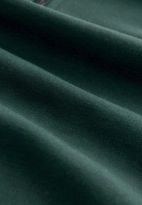 Tmavě zelená látka s jemnou strukturou, obsahující jemné žebrované vzory a jemné záhyby, zachycená v měkkém, přirozeném světle.