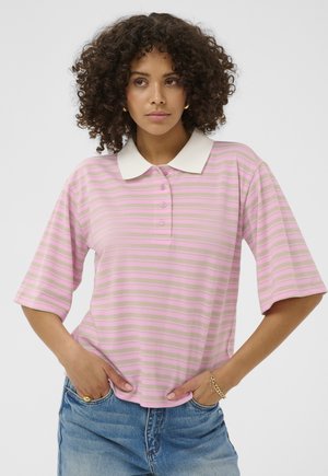 Kurdlike juustega naine, kes kannab roosa-beeži triibulist polosärki ja siniseid teksaseid, käed taskus, ühevärvilise tausta ees.