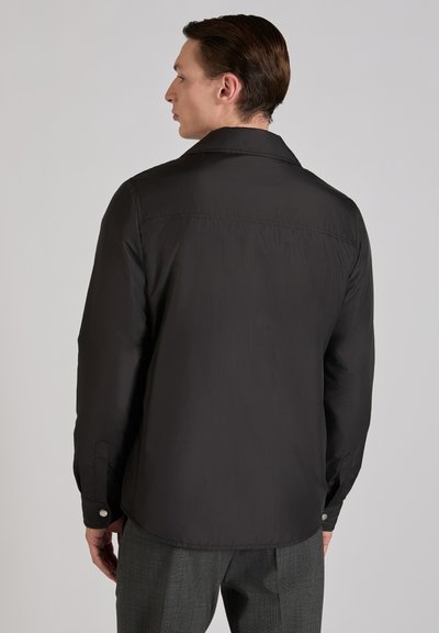 Mężczyzna z zaczesanymi do tyłu włosami, zwrócony bokiem, ubrany w czarną koszulę z długim rękawem i szare spodnie w kratę na jednolitym tle.