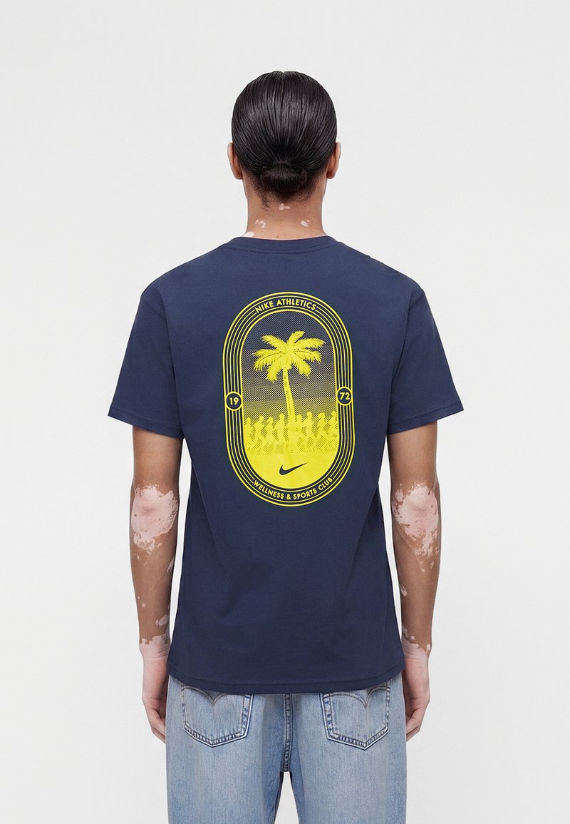 Persona con vitiligine che indossa una maglietta Nike blu navy con disegno giallo di palma e figure che corrono sulla schiena, abbinata a jeans azzurri.