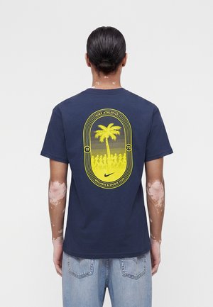 Osoba s vitiligom nosí tmavomodré tričko Nike so žltým dizajnom palmy a bežiacich postáv na chrbte, v kombinácii so svetlomodrými džínsami.