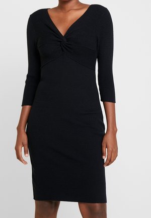 Femme portant une robe noire cintrée avec des manches trois-quarts et un détail de nœud torsadé au col en V.