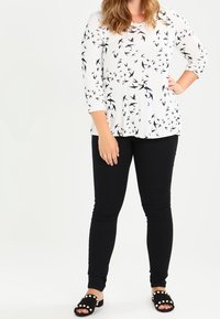 Top bianco con motivo di uccelli neri, maniche a tre quarti; pantaloni neri aderenti; sandali neri con stud bianchi.
