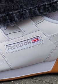Sneaker bianca in pelle con logo Reebok e dettaglio della Union Jack, suola in gomma e texture liscia. Cuciture visibili e design low-top.