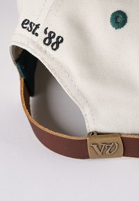 Čepice Cap Tan se zelenými detaily, hnědým koženým páskem, kovovým kováním a vyšitým nápisem "est. '88" na boku. Texturovaný látkový povrch.